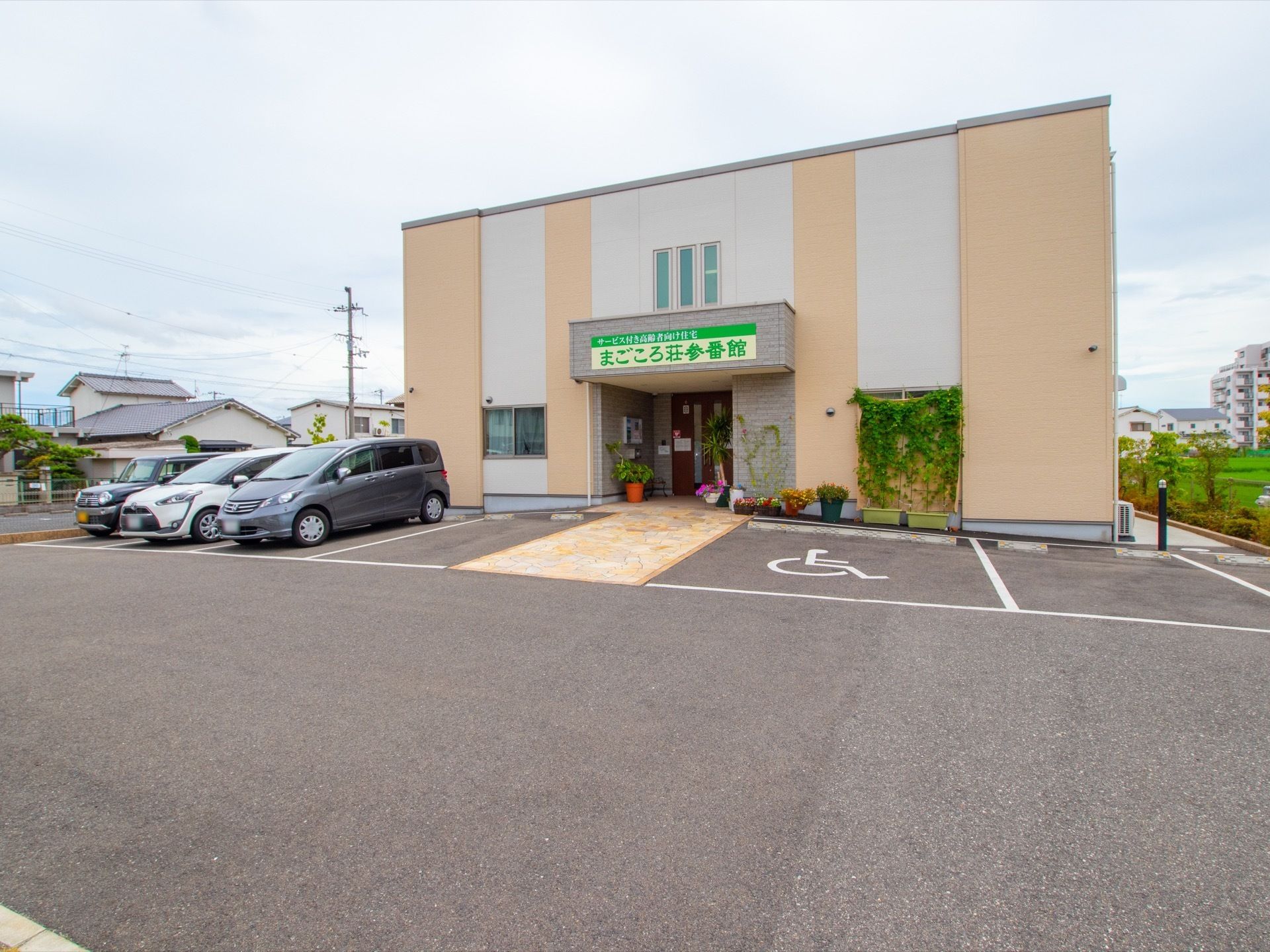 ベージュと白の建物で、玄関のひさしの部分に施設名が書かれている。建物の前に駐車場があり、車椅子専用のスペースも設けられている。