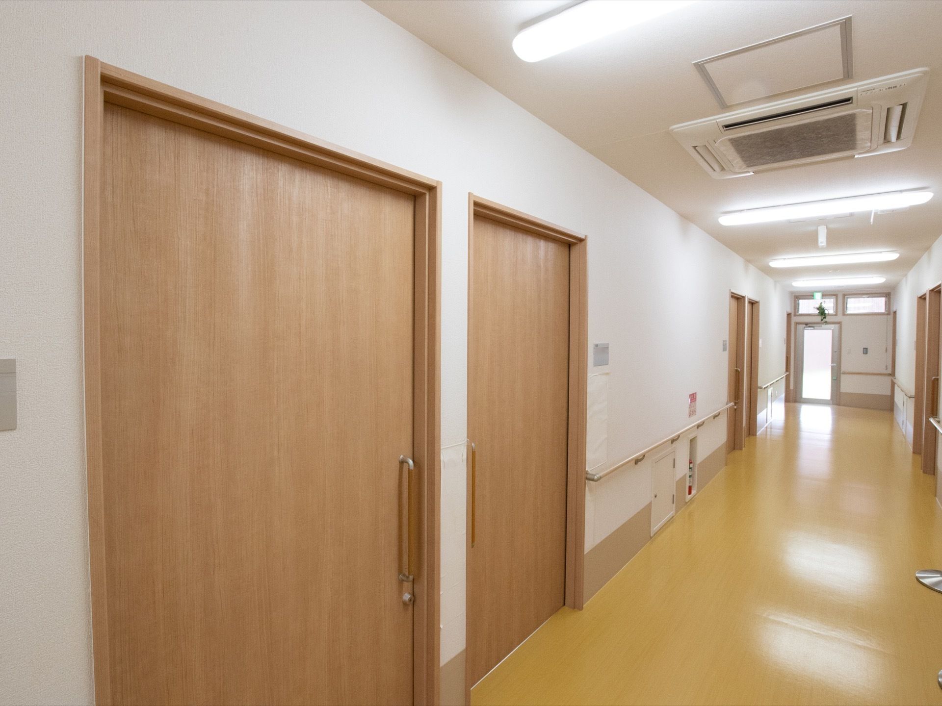 通路には、引き戸になっている木目調の大きなドアが並んでいる。天井には空調設備や、横に長い照明が設置されている。
