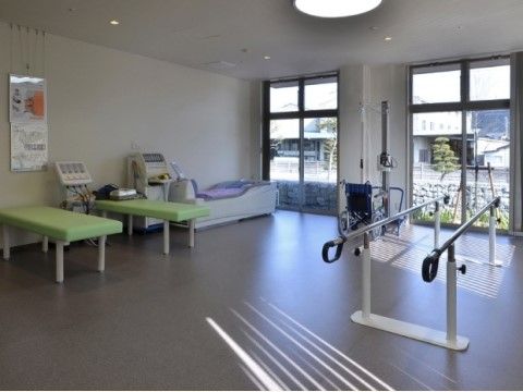 横面にガラス戸が並ぶ広々としたスペースに、歩行器などの機能訓練機器を備えている。壁際に寝台などを置いている。