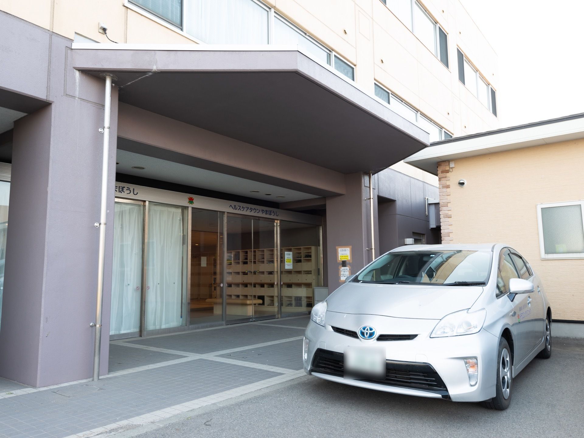 広々とした玄関スペースである。ガラス張りの出入口ドアの上部に施設名を掲げている。前方には大きな屋根があり、１台の車が停まっている。