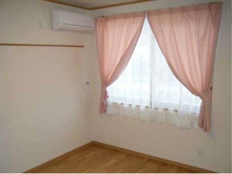 白い壁と明るいブラウンのフローリング。窓があり、薄いピンクのカーテンがついている。エアコンが設置されている。