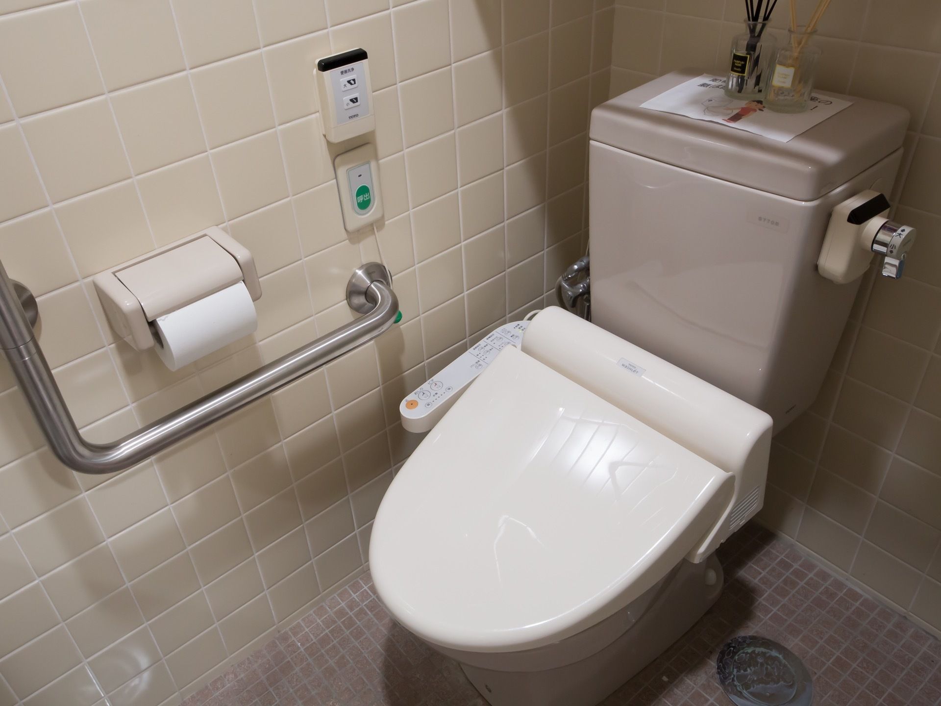 トイレの便座は温水洗浄便座で、近くの壁には銀色の太い手すりが設置されている。施設のスタッフに通じるボタンもついている。
