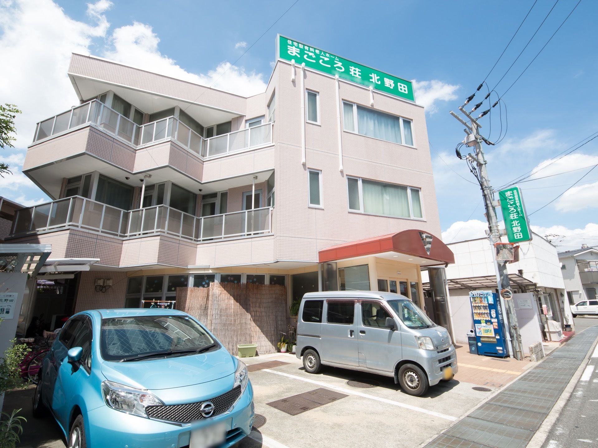 施設の建物の前には、数台分の駐車スペースが設置されている。建物の屋上には大きな緑色の看板があり、施設名が記されている。