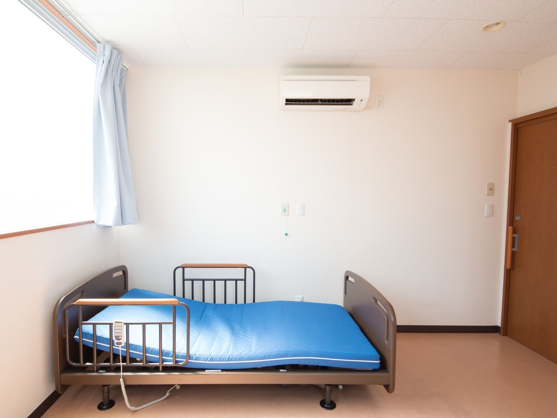 居室のドアは茶色いスライド式のドアである。部屋には青いスプリングマットのベッドが置かれている。エアコンも設置されている。