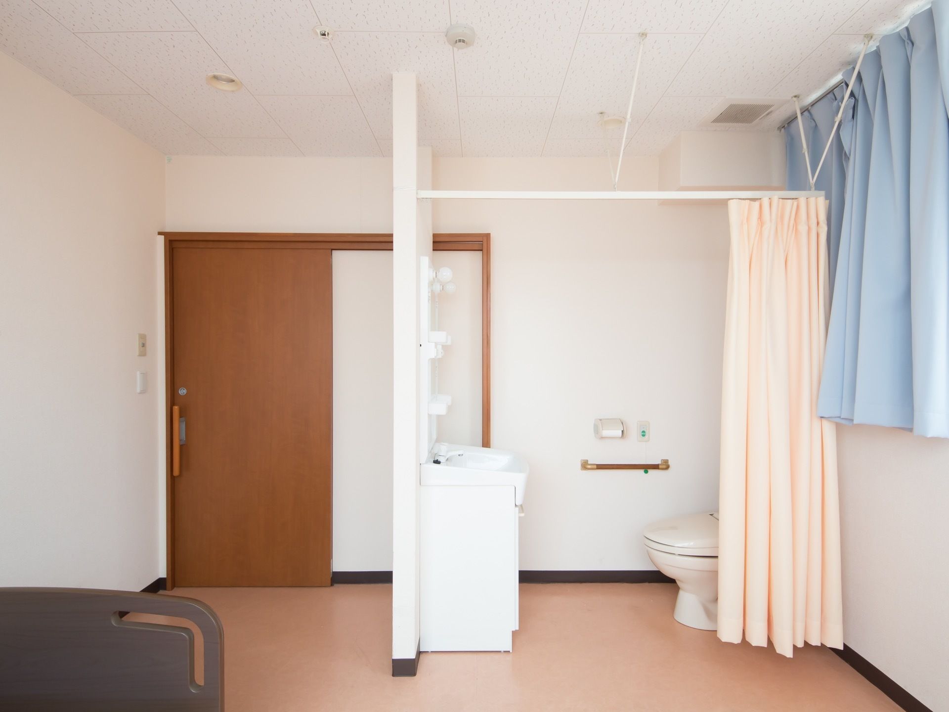 居室内にはトイレと洗面台がある。周囲にはベージュピンク色のシャワーカーテンがついている。トイレの便座のそばには手すりがついている。