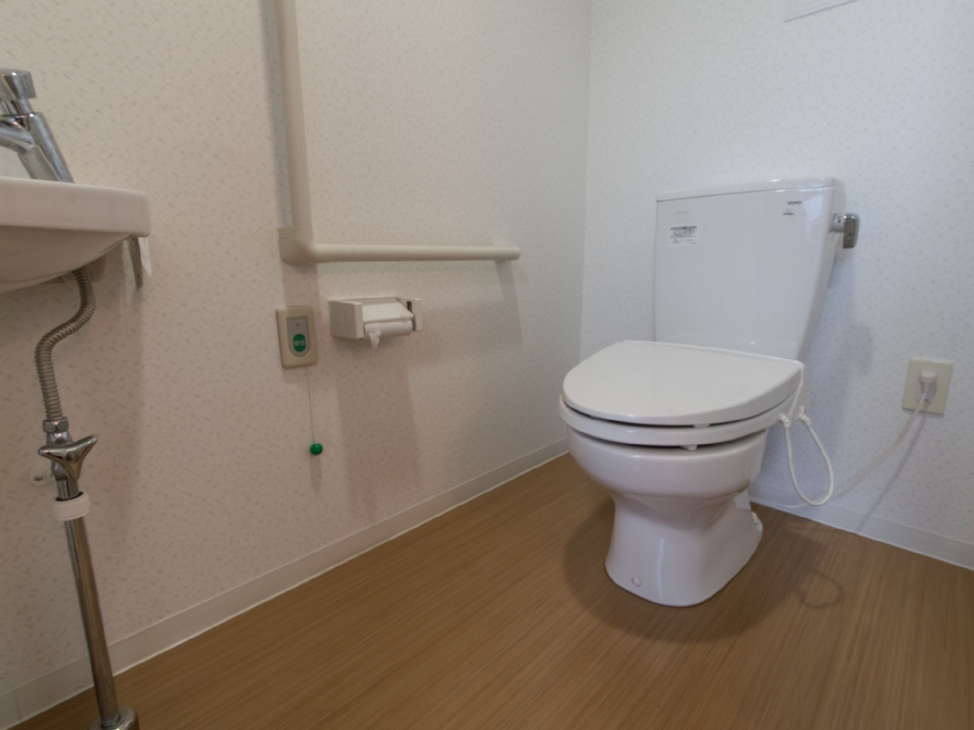 フローリングの床にあるトイレで、洋式である。壁に手すりがあり、そのそばに緊急時の呼び出しベルが設置してある。