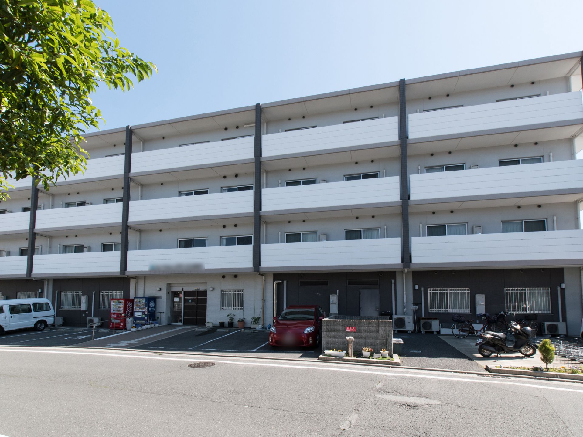 白い外壁の施設で、正面玄関そばに駐車場、駐輪場が設けてある。玄関そばには自動販売機がある。手すりつきスロープがあり、プランターが置いてある。