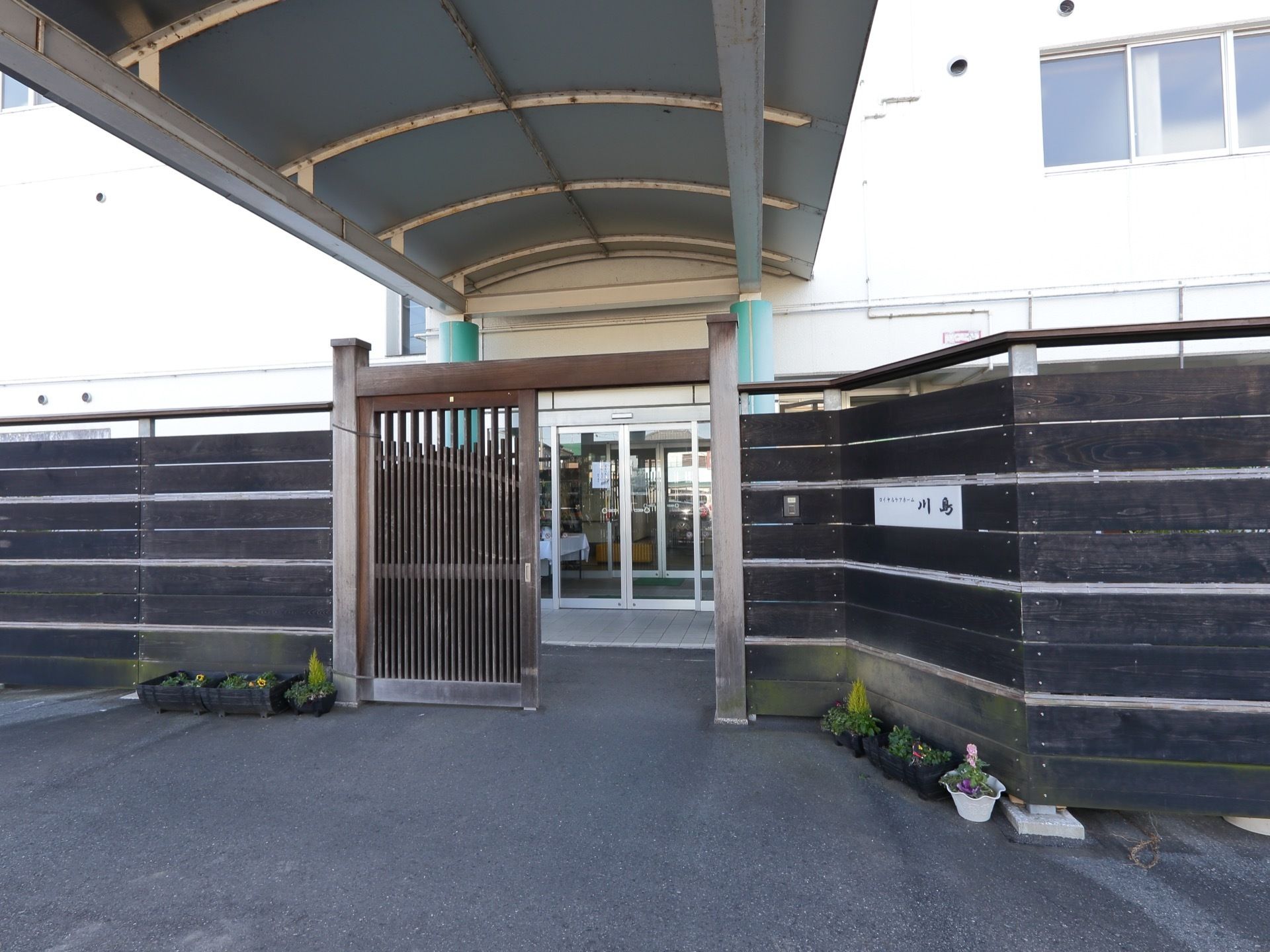 施設の入口は黒い塀で覆われており、シルバーの表札が取りつけられている。入口の門は、木造となっている。