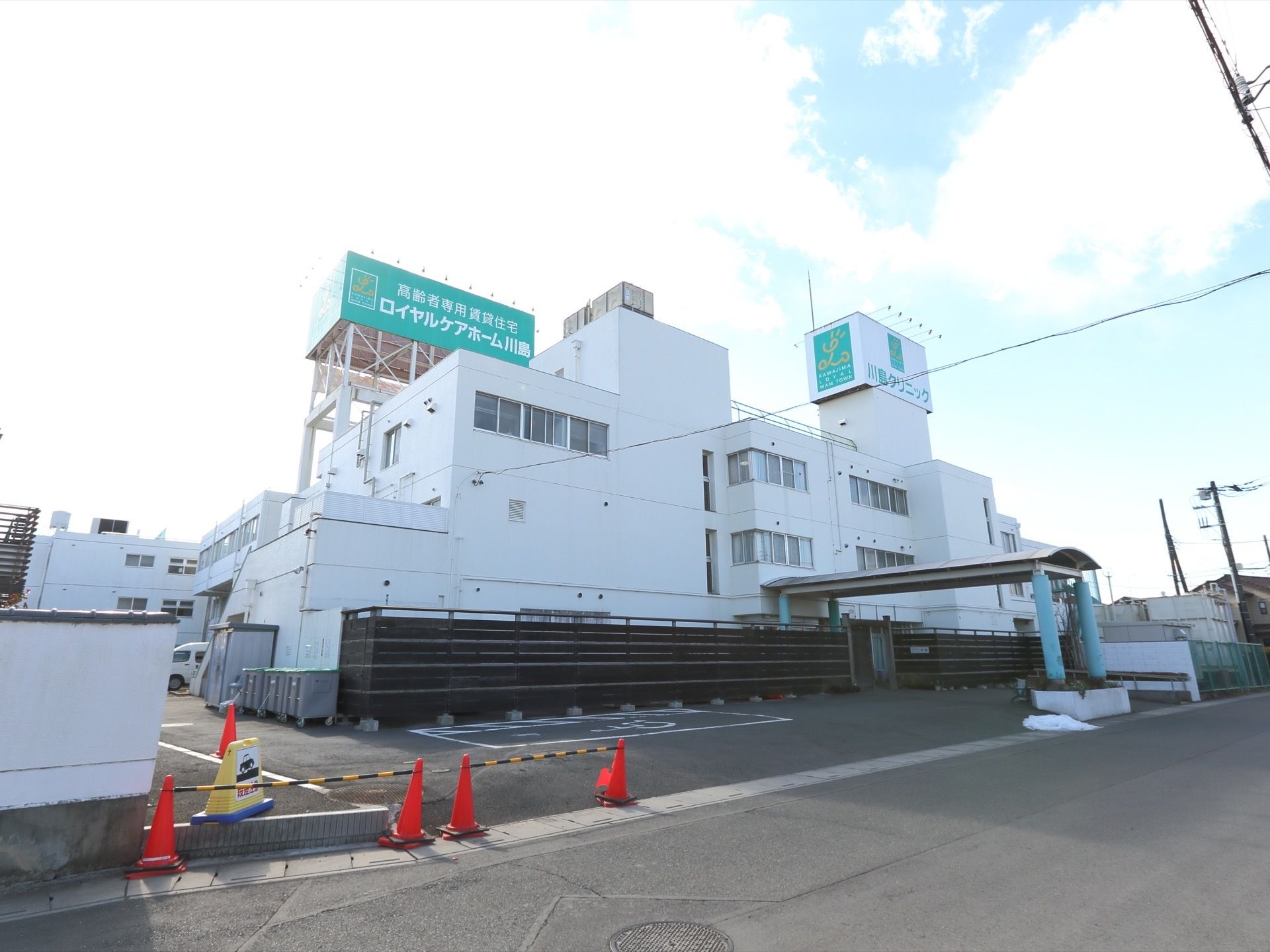 施設は、白い外壁と緑色の看板の取りつけられた外観となっている。施設の周辺には、広い駐車場スペースが確保されている。