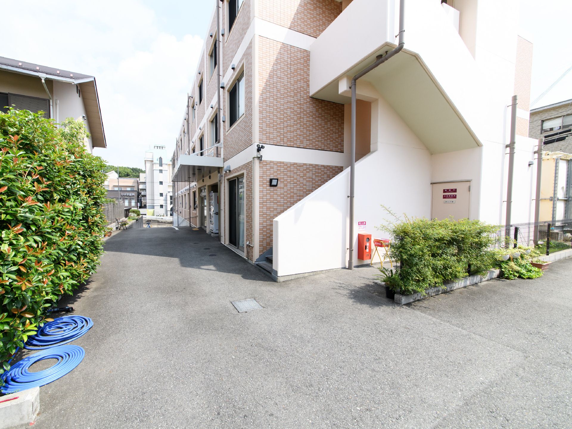 施設の横側は広い道路となっており、民家があって植物が群生している。建物には、非常用の階段が敷設している。