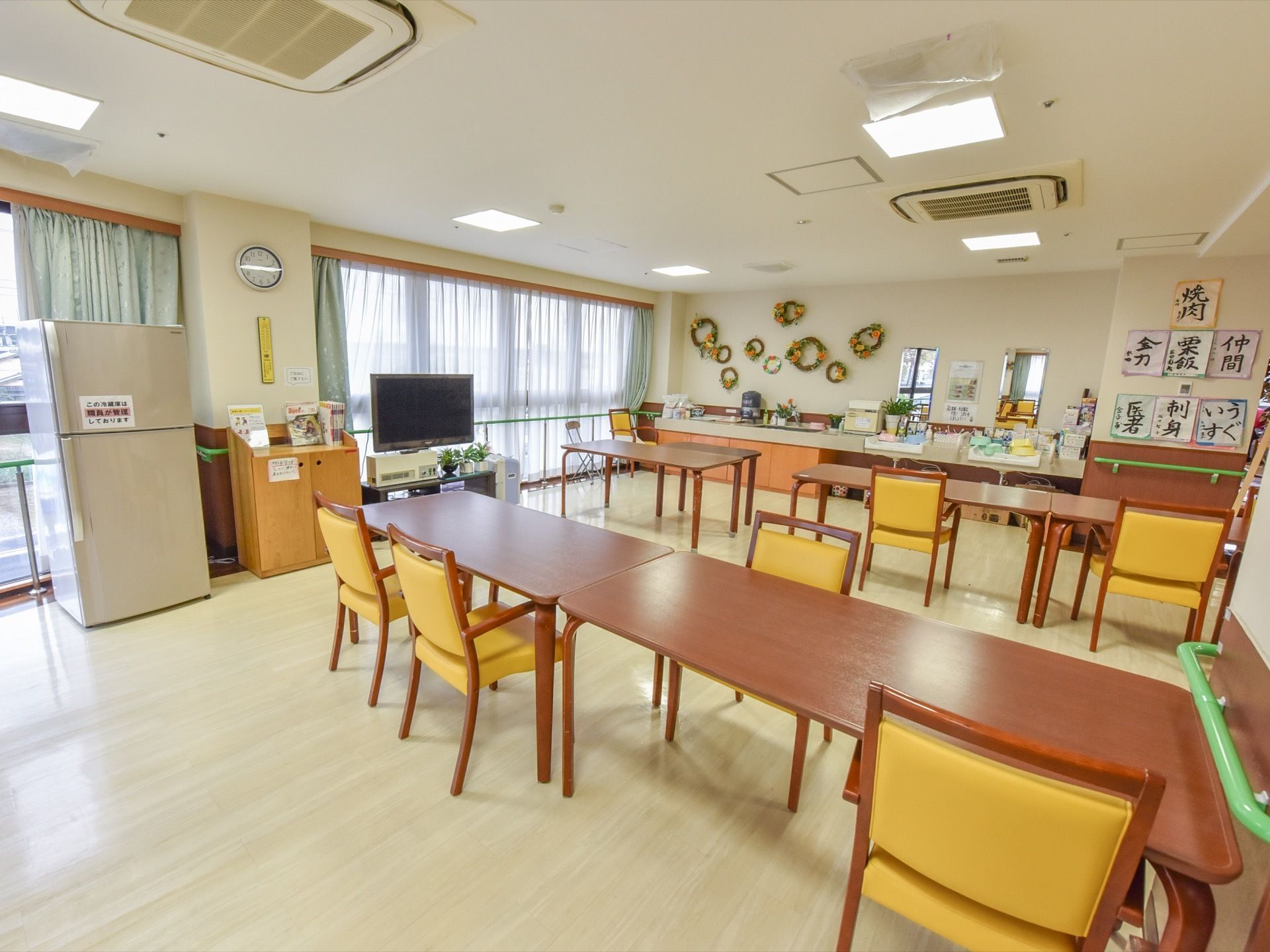 食堂にはテーブルと椅子が整然と並べられている。壁にはリースの他に入居者様による習字作品などが飾られている。