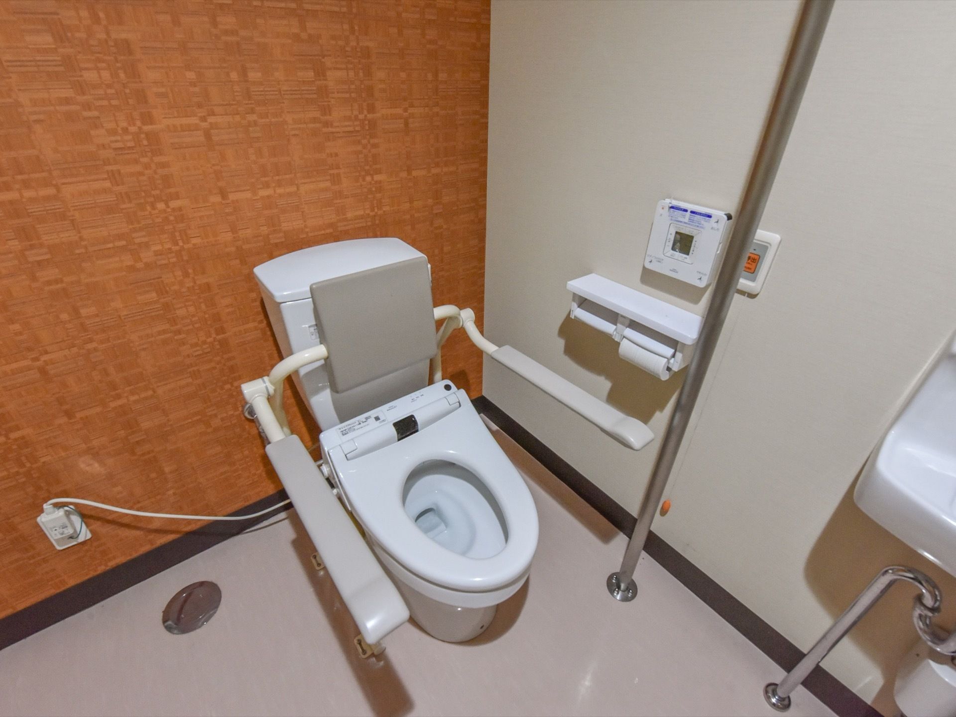 施設利用者様用のトイレ。便座の左手側にはコントローラー類の他、銀色のパイプ状の手すりが床から伸びている。