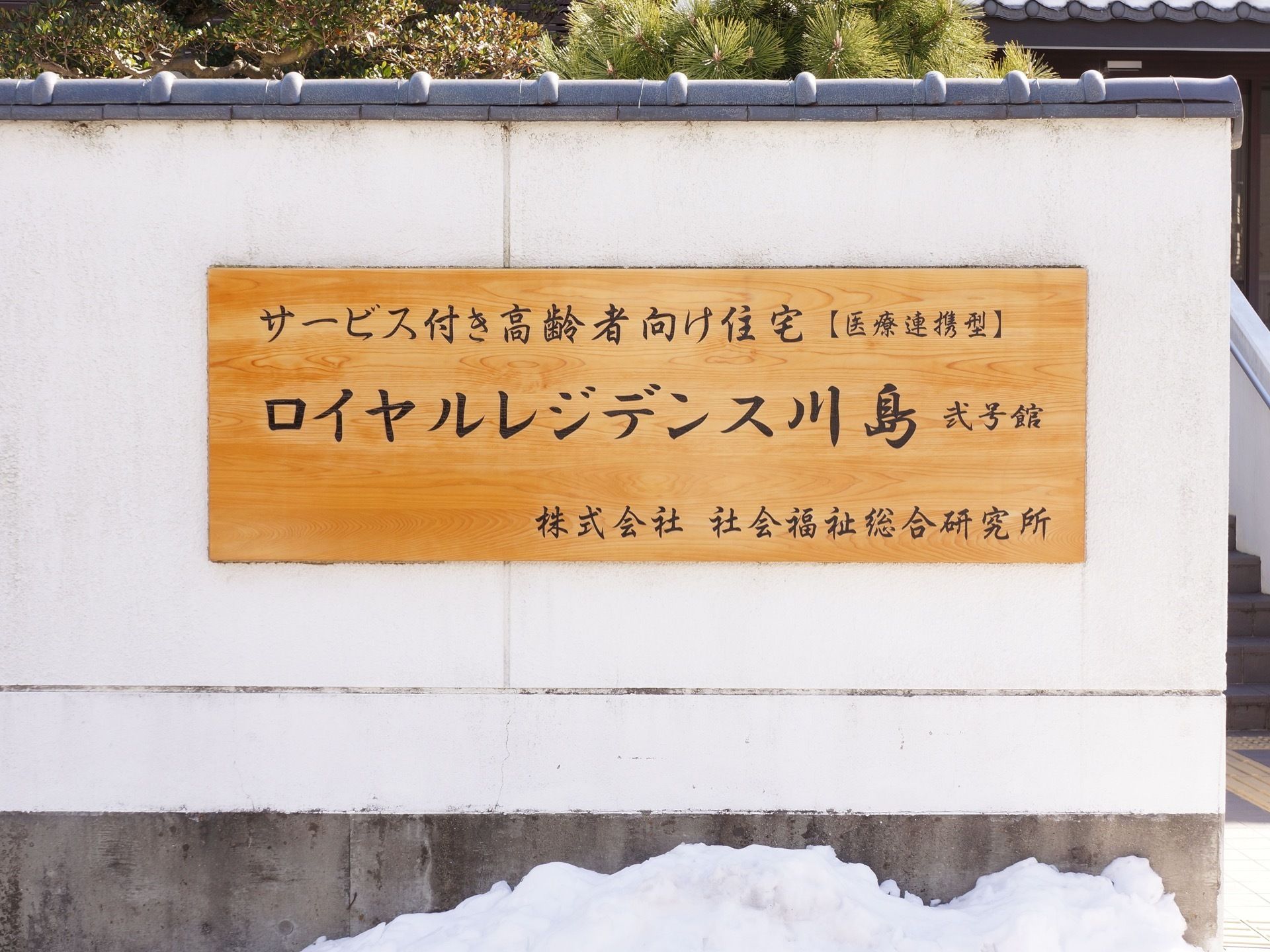 施設の外壁は和風の造りになっている。白い壁には木目調のプレートが貼られ、行書体で施設名、施設の種類、運営会社名が書かれている。