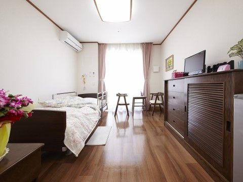 窓際にテーブルと椅子、ベッドや大きなタンスが設置されている花のある居室
