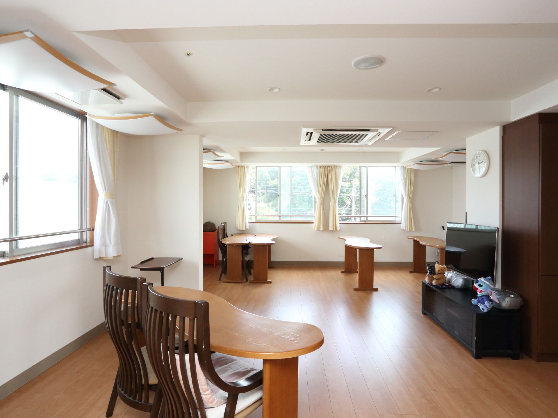 手すりのついた窓がある、ウッドフロアの部屋である。壁掛け時計の下にテレビが置いてあり、リモコンが床に置いてある。