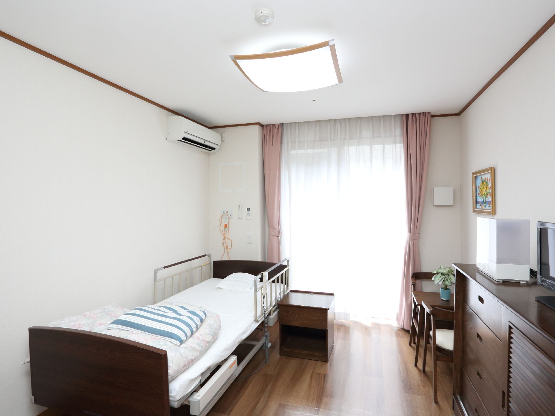 白い壁にウッディなフロアの部屋で、木製の机が置いてある。窓に手すりがついていて、白いカーテンが下がっている。
