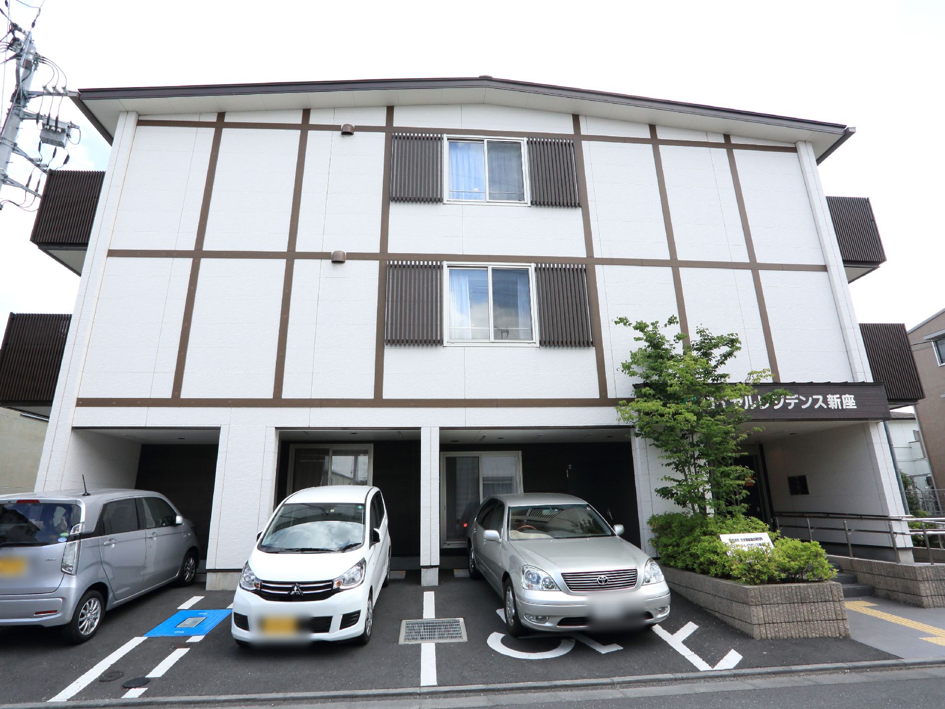 白い外壁で、茶色のベランダフェンスがある施設で、玄関そばには手すりが設置してある階段とスロープが設けてある。