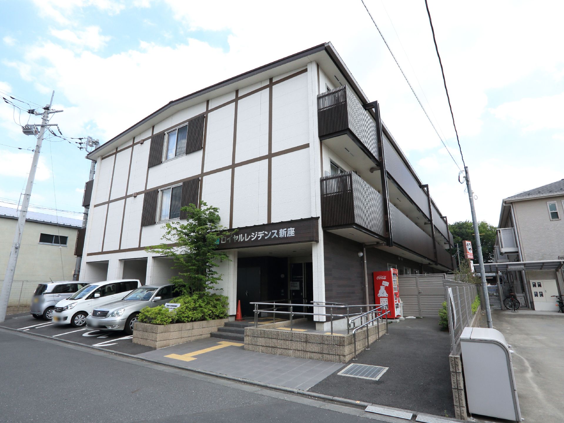 玄関の横に自動販売機がある。隣との境界には、メッシュフェンスがある。玄関のポーチの屋根部分に施設名が大きく掲げてある。