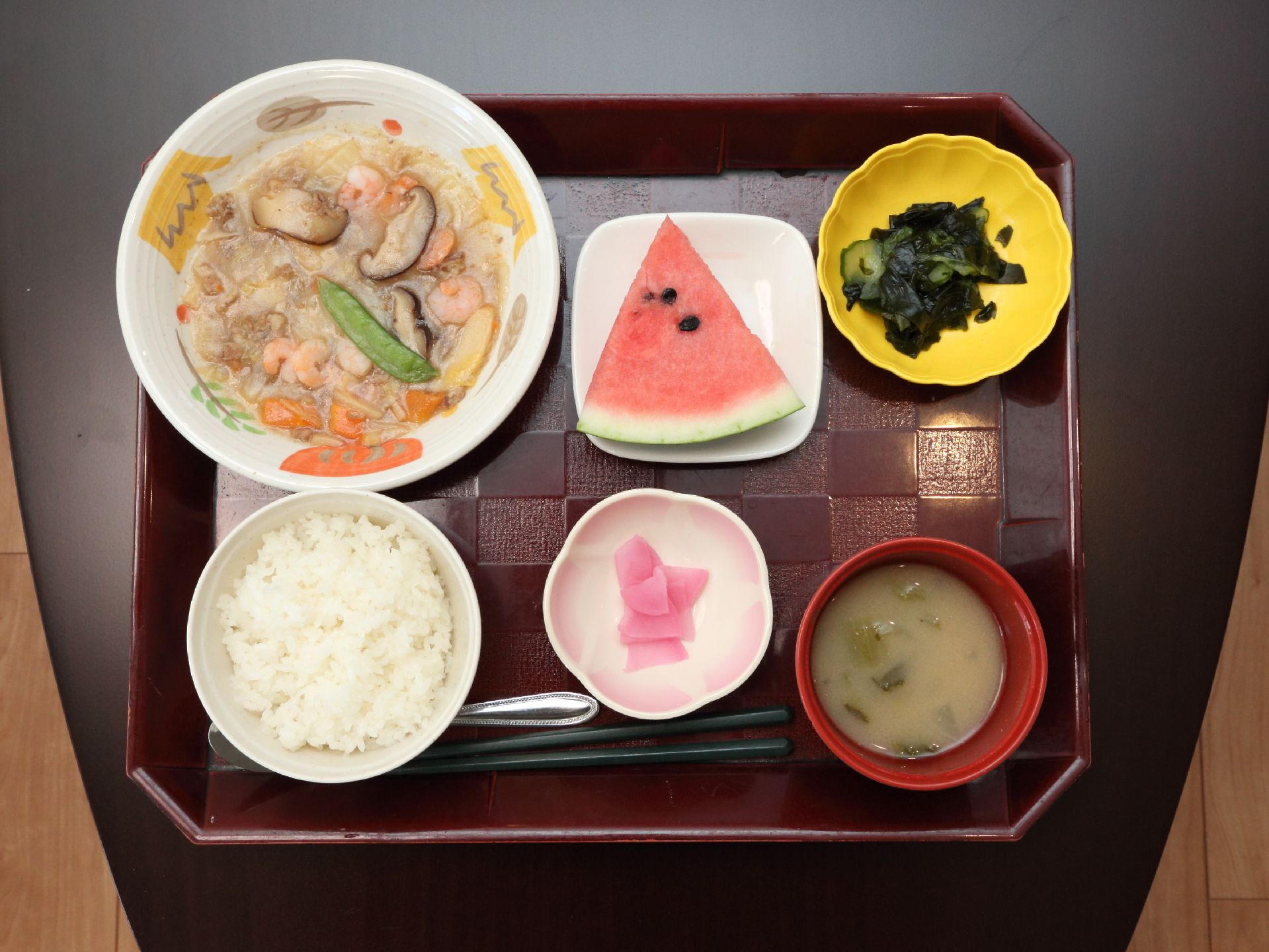 こげ茶色のテーブルの上に、ワインレッドカラーのお盆がおかれ、その上に食事が盛られている。緑の橋とスプーンがある。