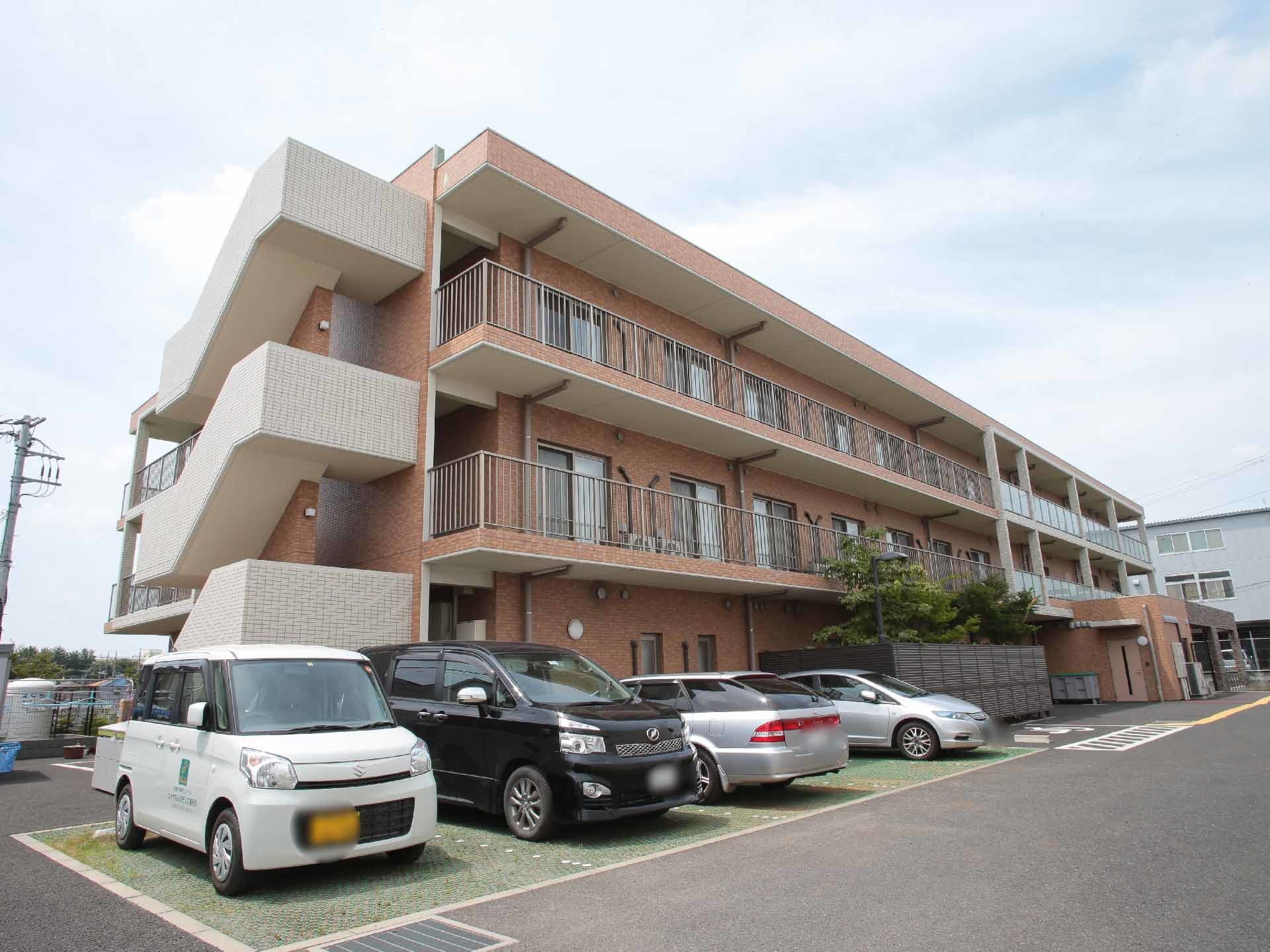 広々とした敷地に建てられている3階建ての施設の外観。サーモンピンクの外壁が明るい雰囲気がある。建物の前には駐車スペースが設けられている。
