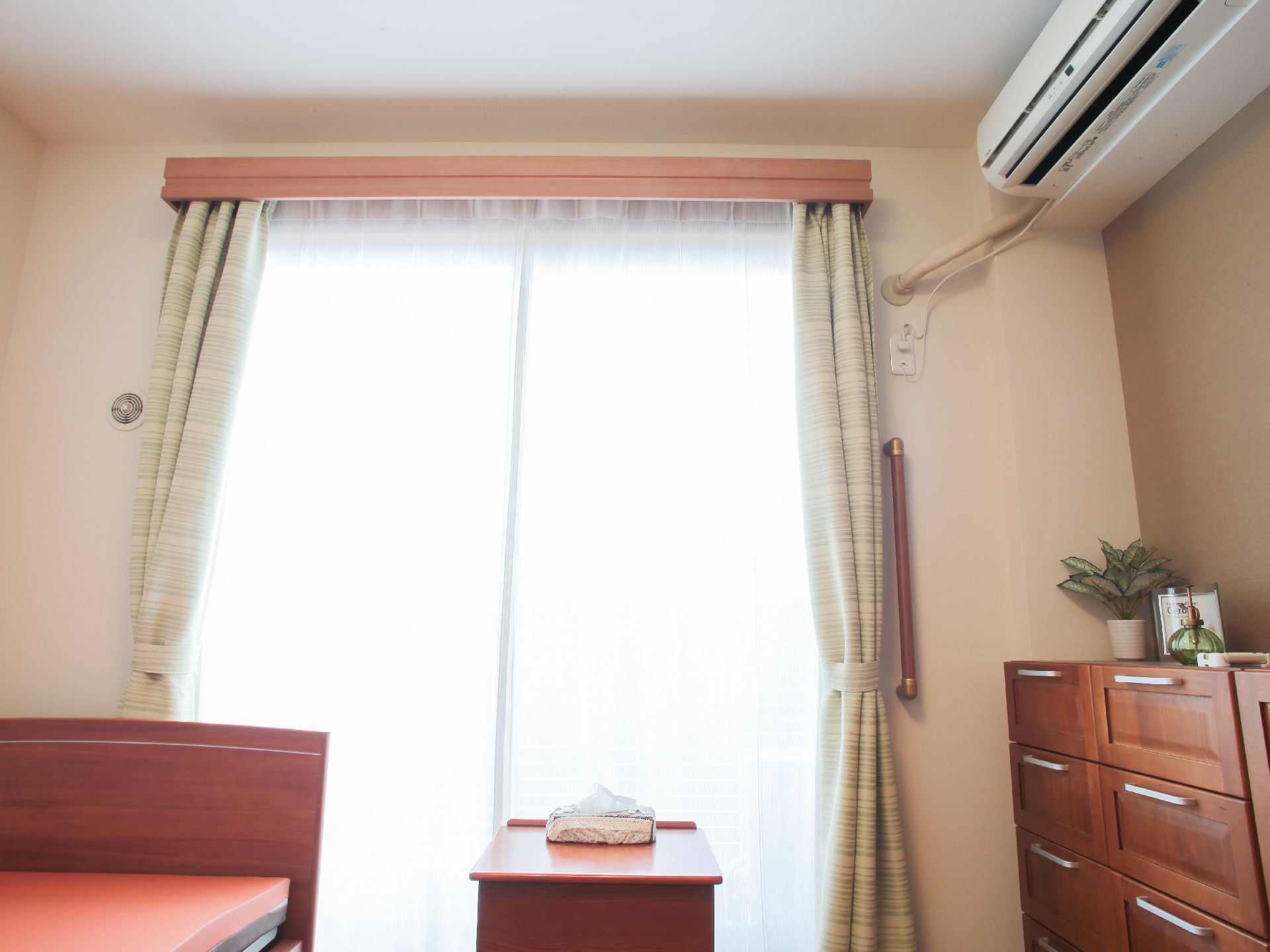 大きな窓から入る光でとても明るい部屋となっている。ベッドや収納がある。エアコンが設置されており身体への配慮もされている。