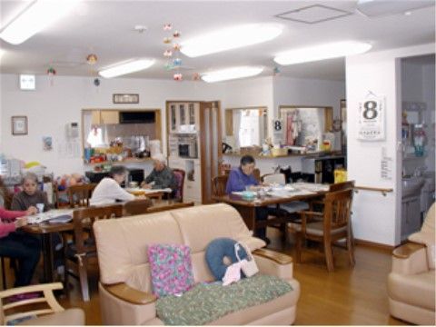 手前にソファがあり、奥にはダイニングセットが３つ置かれている。入居者様が本を読んだりお話をなさっている。奥には台所がある。