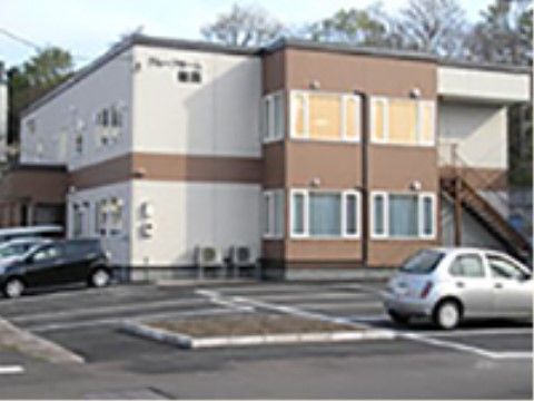 茶色と白で統一されたシンプルなデザインの施設。緑に囲まれた施設の前には、十数台停めることのできる駐車場が設置されている。