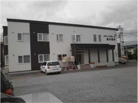 駐車場から見た建物の全景で、二階建ての建物。エントランスには階段とスロープがあり、屋根が張り出している。