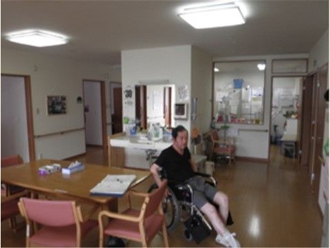 ダイニングテーブルの横には車椅子をご利用の方がいらっしゃる。左手奥には個室が並んでいる。奥はガラス張りでキッチンの様子がわかる。