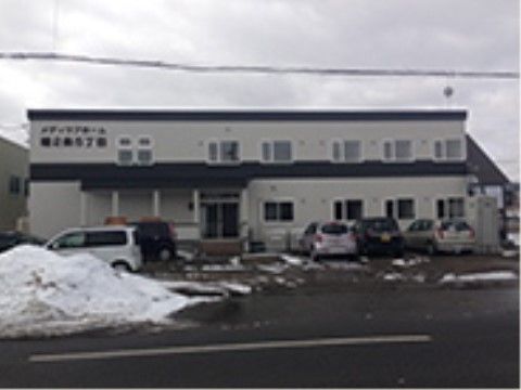 白色の２階建ての建物で、施設名が外壁に掲示されている。建物の前には駐車場が設けられており、車が数台駐車されている。