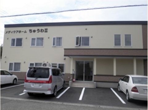 ベージュの外壁が落ち着いた雰囲気の施設の外観。建物は2階建てで、建物の前には駐車スペースがが設けられている。