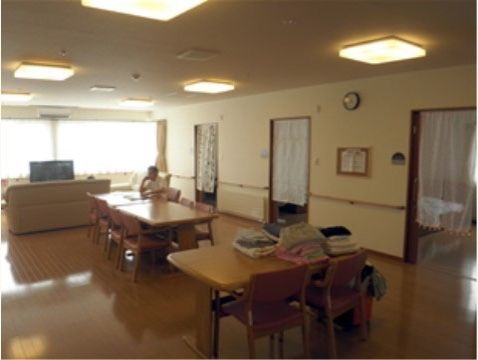 居室から扉一枚でつながっているリビング。木製のダイニングテーブルセットが置かれている。1名の入居者様がテーブルについている。