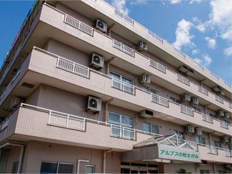 ベージュが基調になった団地風の建物の正面。各部屋のベランダが見えており、壁上部に室外機が設置されている。