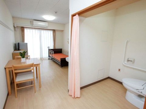 広いフローリングの部屋。手前にフタが閉まっているトイレがあり、ピンクのカーテンが仕切り代わりになっている。