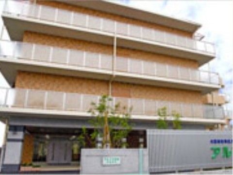 左側に施設の入り口があり、両開きのドアが見える。そばには大きな木が数本生えており、その手前には白い塀がある。