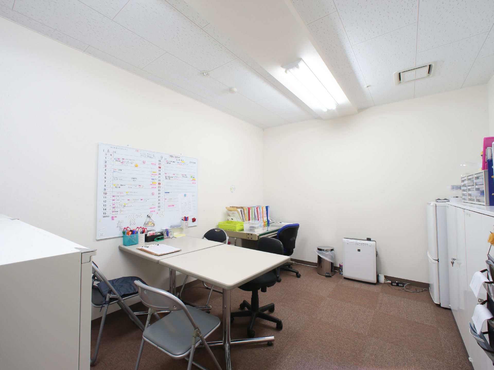 白いテーブルが二つ並べて置いてあり、横の壁にスケジュールボードが取り付けてある。デスクとチェアがが角にありファイルがたくさん置いてある。