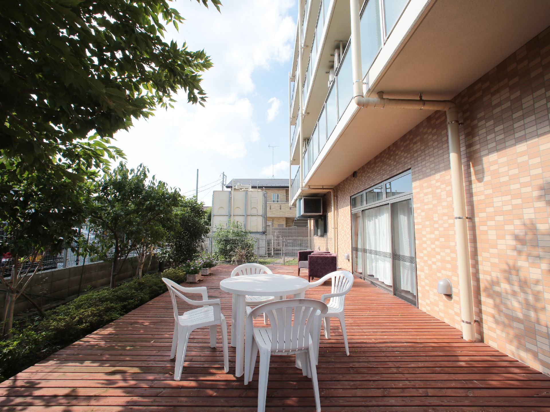 ウッドデッキがあり、白いフェンスで囲まれている。プランターが左手奥に見える。低木が植栽してある。白いガーデンテーブルとチェアがある。