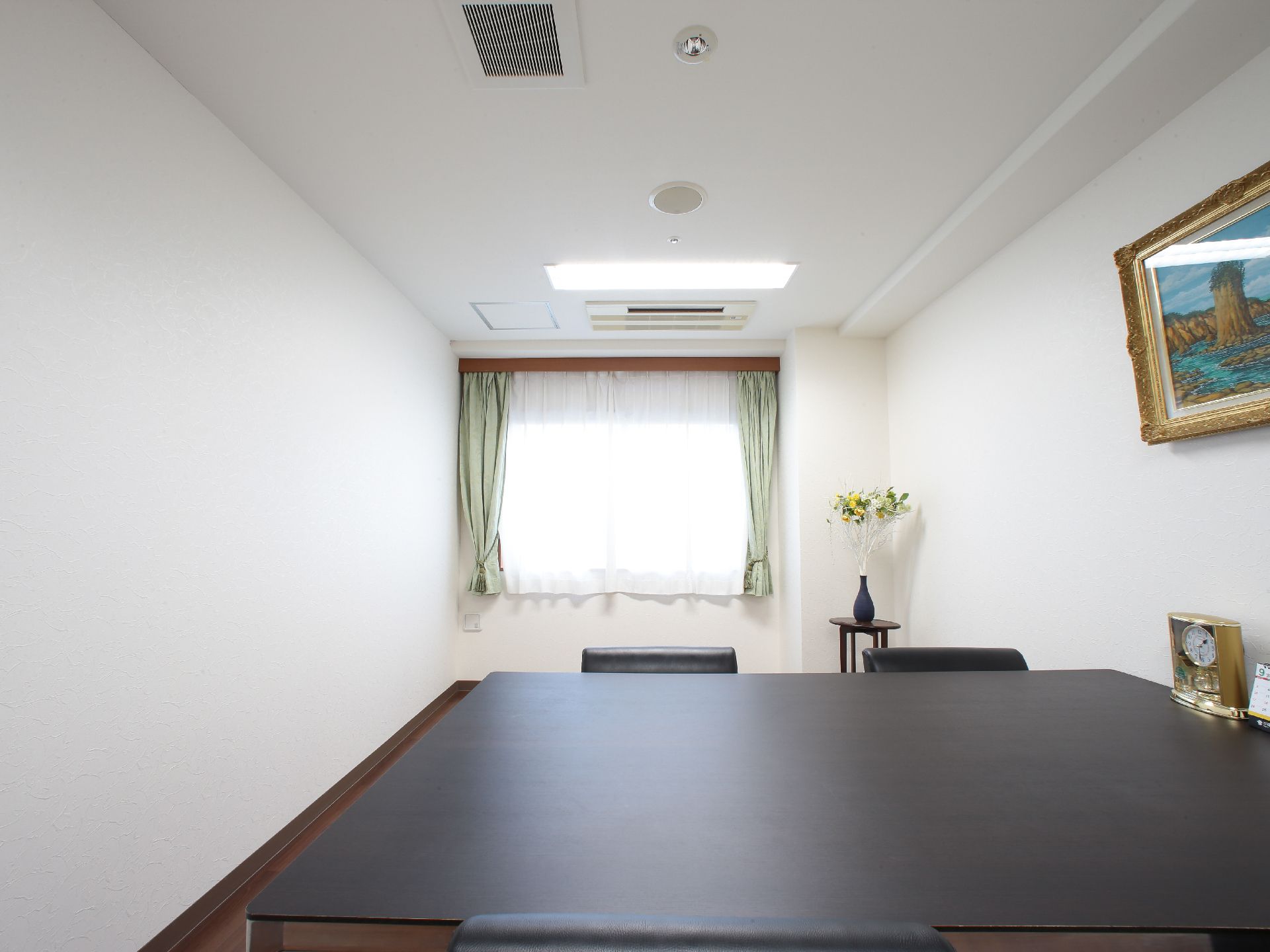 部屋の真ん中あたりに黒い四角いテーブルがあり、椅子が並べてある。右手の壁に絵画が飾ってある。机上に置時計がある。