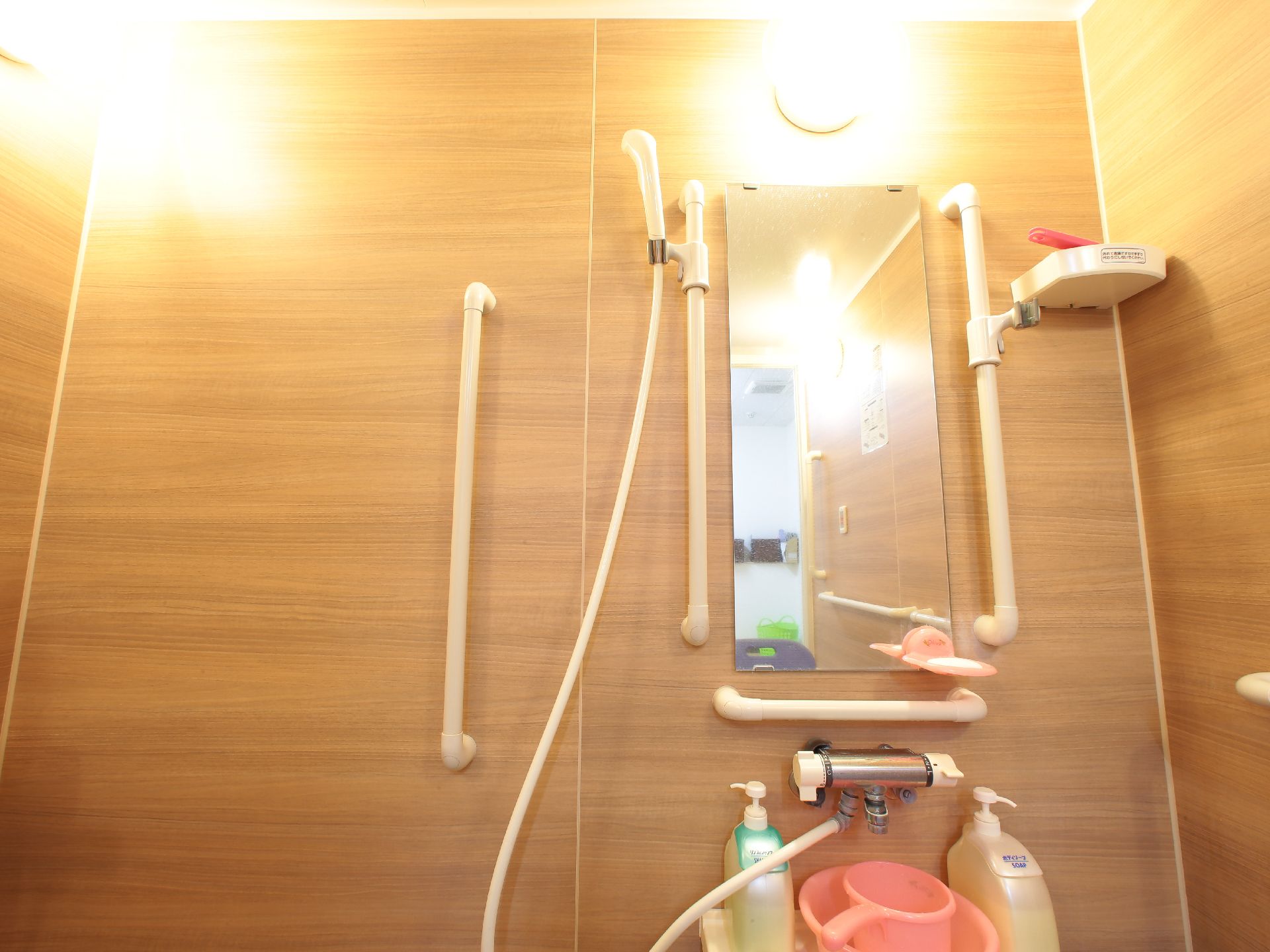 壁にシャワーフックが取り付けてある。鏡を囲うように手すりが取り付けてある。シャンプーやリンスなどのボトルが置いてある。
