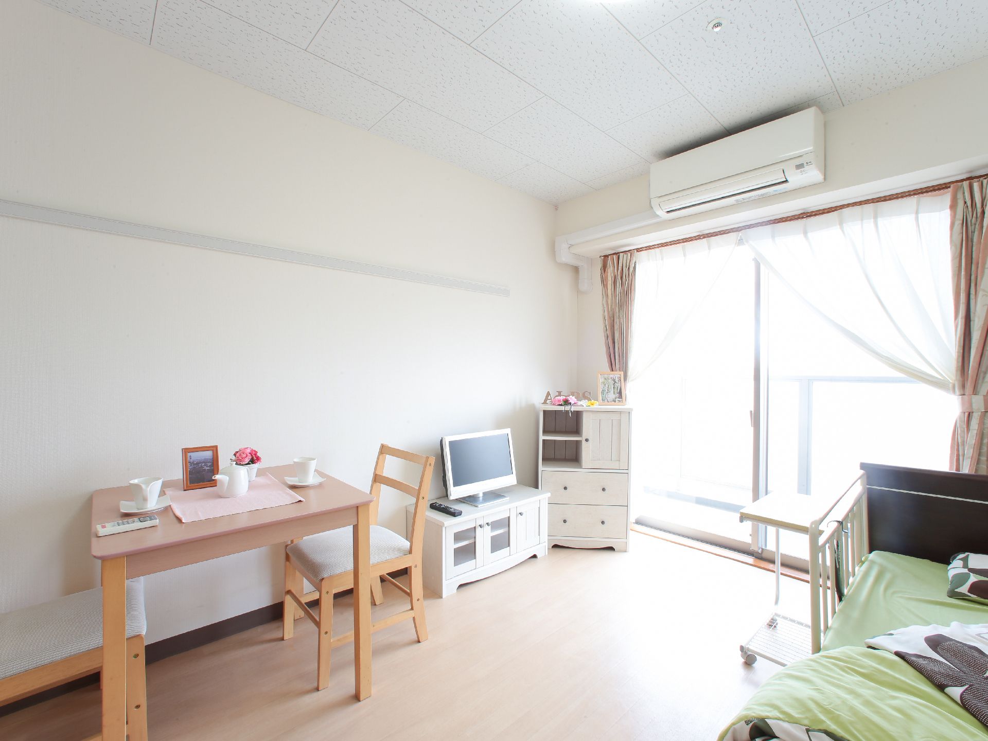 左手に白い収納棚が在り、天板にフォトたてがある。ローボードにテレビが置いてある。四角いテーブルがあり、マグカップとソーサーが二客ある。