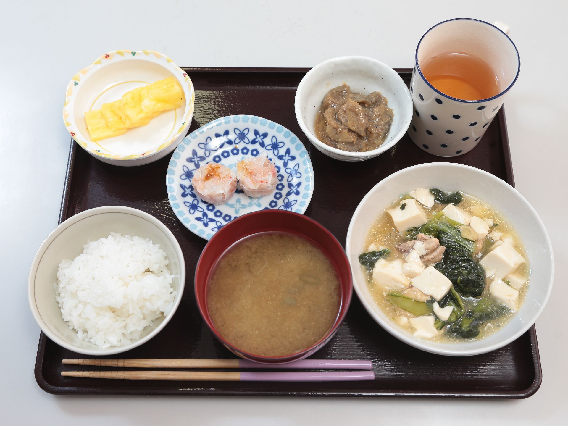 黒色のお盆の上にごはん、味噌汁のほかに、惣菜が三品お皿に盛り付けて置いてある。箸が一膳ついている。湯飲みがある。