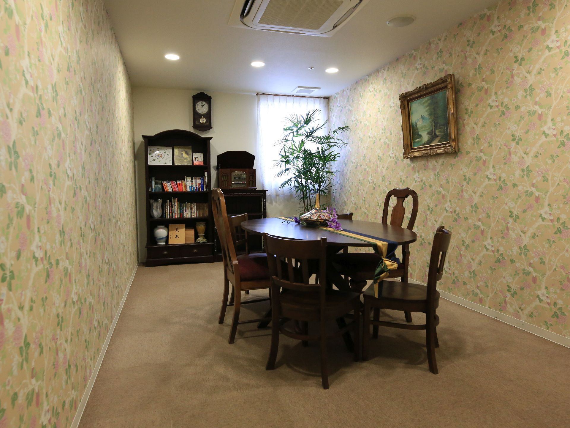 植物の模様の壁紙に囲まれた小部屋。ダークブラウンのテーブル、椅子、棚が置かれている。壁には絵と時計が掛けられている。