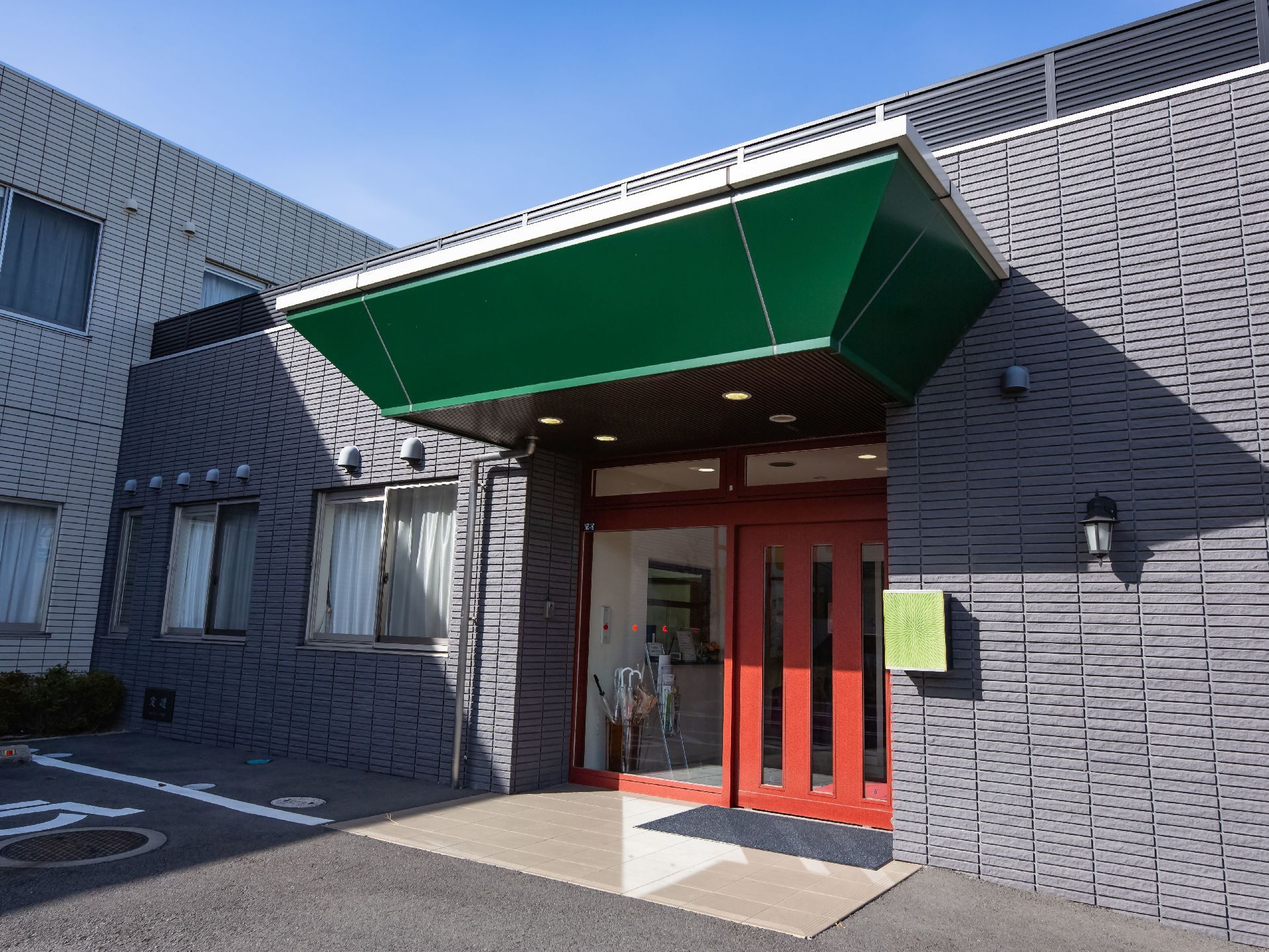 グレーのタイルの壁の施設の玄関がある。赤茶色の木の自動ドアがあり、床には段差がなく玄関マットが敷かれている。