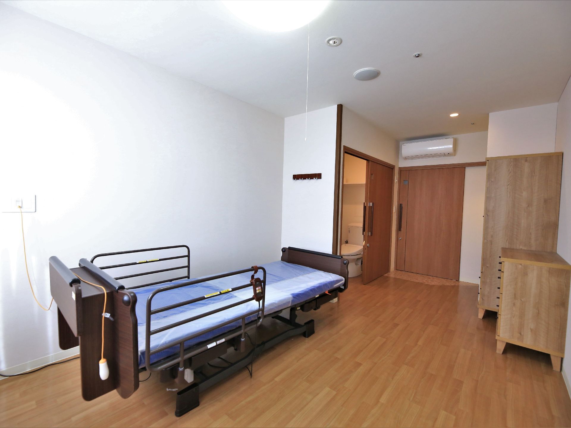フローリングの居室がある。木の素材を使った棚が置かれている。入口付近にはトイレと、壁際には介護用ベッドが置かれている。
