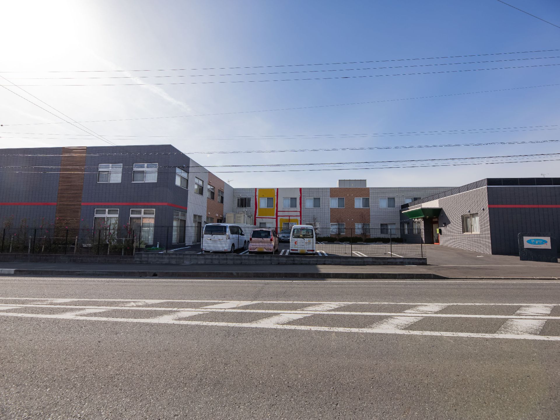 大きな道路に面した二階建ての大きな建物がある。建物の壁面は複数の色で塗装されている。前面は広い駐車場になっている。