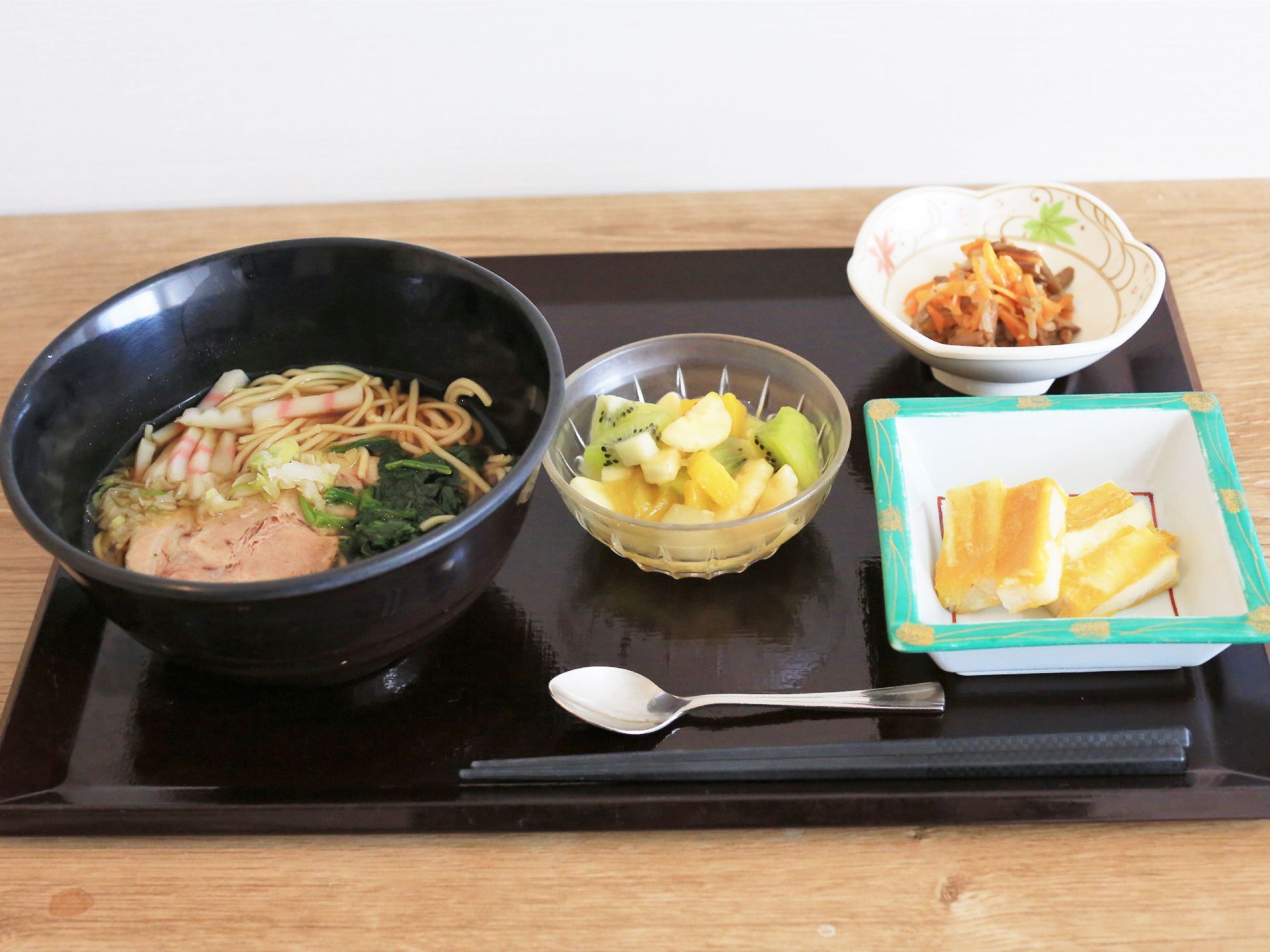 黒塗りのお盆の上に置かれた食事。黒いどんぶりに入った蕎麦、押し寿司、煮物、フルーツポンチが置かれている。