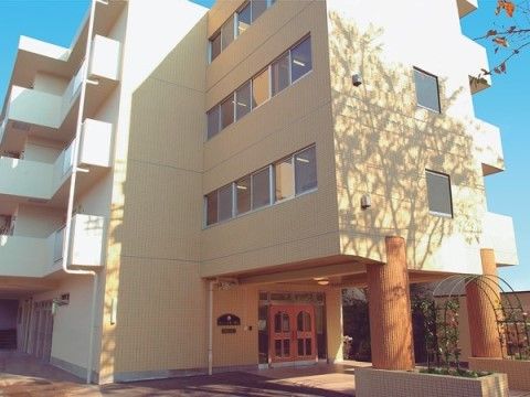 「サニーライフ福生」の外観。住宅と自然に佇むライトベーシュの４階建てのマンション型施設になっている。