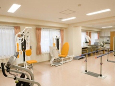 窓がある明るい室内に、トレーニングマシーンが３台と平行棒がある。奥には机と椅子が並び、机の上には胡蝶蘭の鉢がある。