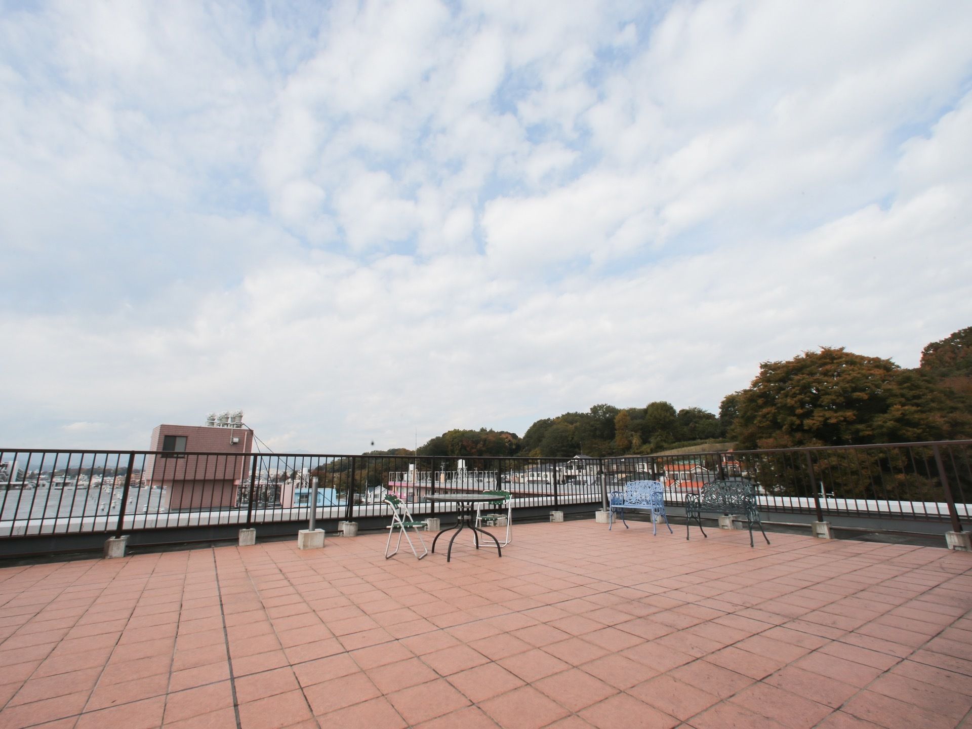 屋上にはイスやテーブル、ベンチといったものが置かれている。空は雲がかかっており、遠くには緑の木々が見える。