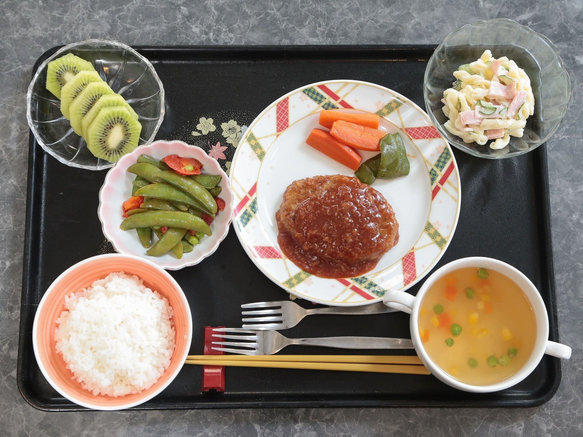 花模様の黒いお盆の上にはさまざまな食事が載っている。手前には２つのフォークと箸置きに置かれた箸が添えられている。